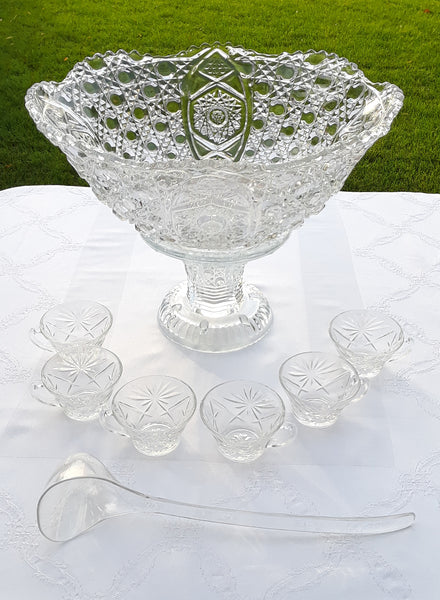 A clear glass punch bowl with a button pattern, accompanied by a clear plastic ladle and a set of matching cups displayed on a white tablecloth.