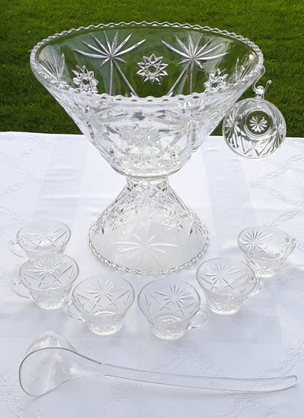A clear glass punch bowl with a star pattern, accompanied by a clear plastic ladle and a set of matching cups displayed on a white tablecloth.